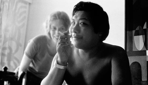 Chogyam Trungpa Rinpoche sits shirtless, with cigarette in hand, and woman in background.