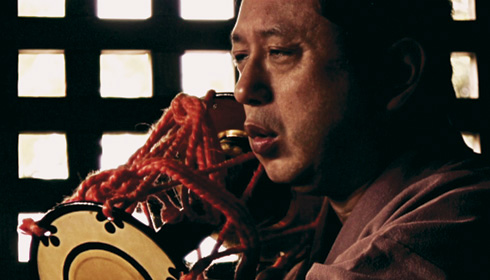 [Japanese man vocalizes while holding instrument tied with red cord]

