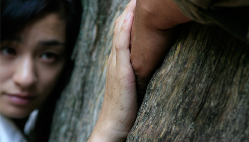 [leaning on a tree trunk, a Japanese woman's face gazes at her hand clasping a man's hand]