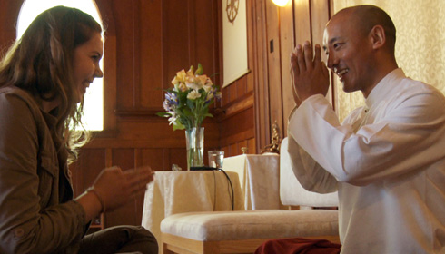 [seated female student is greeted in gasho by seated Anam Thubten in light filled room]